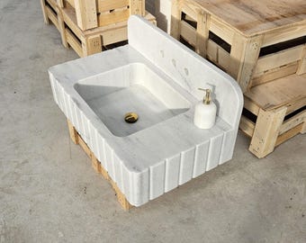 May include: A white marble wall-mounted sink featuring a rectangular basin, a high backsplash with three pre-drilled faucet holes, and a fluted front design. A gold-toned drain cover sits in the basin, and a white soap dispenser rests on the deck.