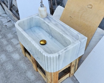 May include: A rectangular white marble farmhouse sink featuring a fluted exterior design. The basin includes a polished gold-tone drain cover. A matching marble soap dispenser with a silver-tone pump is positioned on the flat rear ledge of the sink.