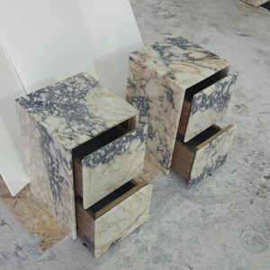 May include: Two marble nightstands with open drawers. The nightstands are rectangular with a cream and gray marbled pattern. The drawers are open, revealing a wooden interior. The nightstands are on a concrete surface.