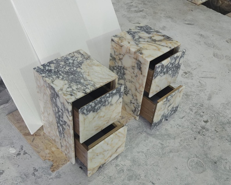 May include: Two marble nightstands with open drawers. The nightstands are rectangular with a cream and gray marbled pattern. The drawers are open, revealing a wooden interior. The nightstands are on a concrete surface.