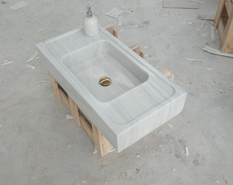 May include: A rectangular white marble sink featuring a smooth basin with a gold-toned drain plug. A white ceramic soap dispenser sits on the flat edge of the sink. The sink is supported by a light-coloured wooden frame structure.