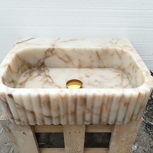 May include: A rectangular marble sink with a scalloped edge. The sink is a creamy white color with brown veining. A gold-colored drain is visible in the center of the basin. The sink is sitting on a wooden pallet.