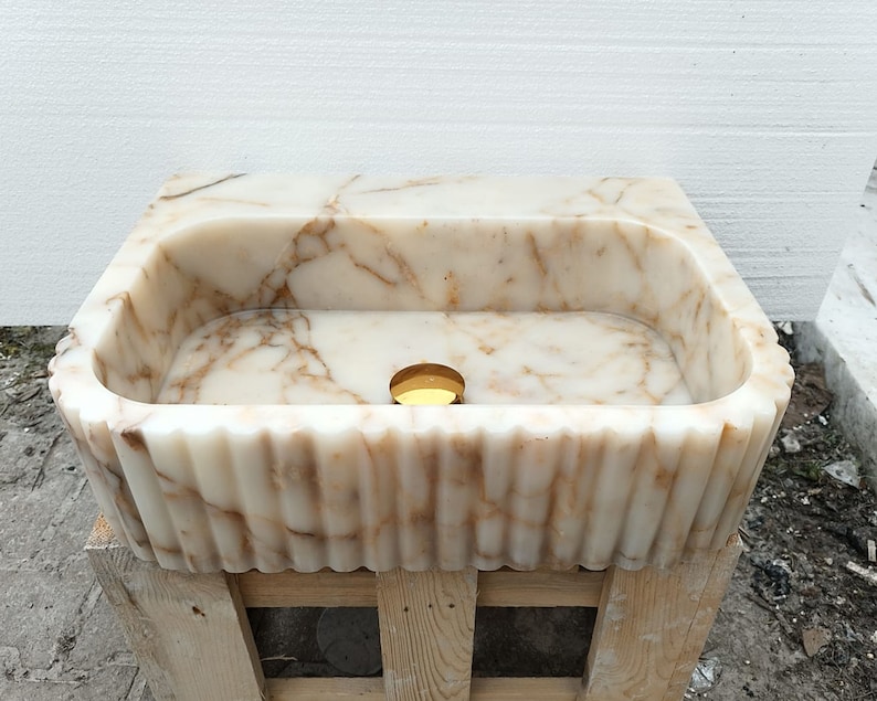 May include: A rectangular marble sink with a scalloped edge. The sink is a creamy white color with brown veining. A gold-colored drain is visible in the center of the basin. The sink is sitting on a wooden pallet.