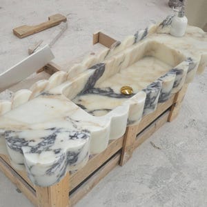 Puede incluir: Un lavabo rectangular de m&aacute;rmol con un dise&ntilde;o de borde festoneado y vetas blancas y grises. El cuenco incluye una tapa de desag&uuml;e dorada centrada. Una peque&ntilde;a botella blanca se asienta sobre la superficie plana de la estructura de piedra.