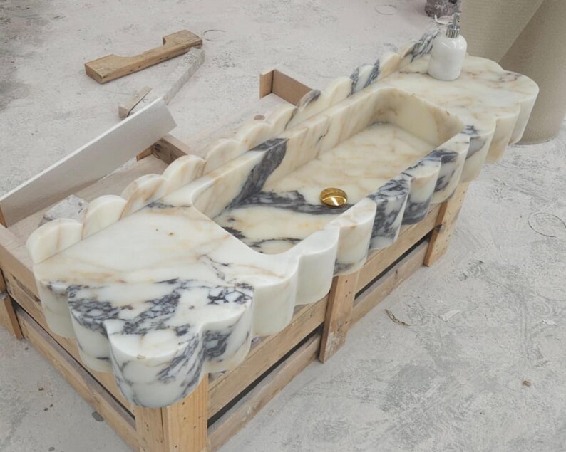 Puede incluir: Un lavabo rectangular de m&aacute;rmol con un dise&ntilde;o de borde festoneado y vetas blancas y grises. El cuenco incluye una tapa de desag&uuml;e dorada centrada. Una peque&ntilde;a botella blanca se asienta sobre la superficie plana de la estructura de piedra.