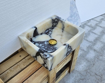 May include: A square bathroom sink carved from polished white marble with prominent black veining throughout the stone. The basin features a smooth, rounded interior and a centered, circular gold-toned metal drain cover, showcasing natural stone patterns.