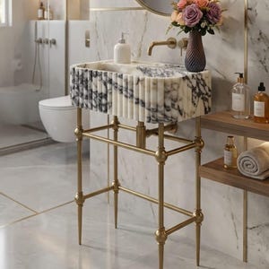 Puede incluir: Un lavabo de mármol con una estructura metálica dorada. El lavabo tiene una pila rectangular con un patrón en blanco y negro. Un jarrón con flores y un dispensador de jabón están sobre el lavabo. El baño tiene una pared y un suelo de mármol.
