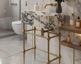 May include: A marble vanity sink with a gold-colored metal frame. The sink has a rectangular basin with a black and white pattern. A vase of flowers and a soap dispenser sit on the sink. The bathroom has a marble wall and floor.