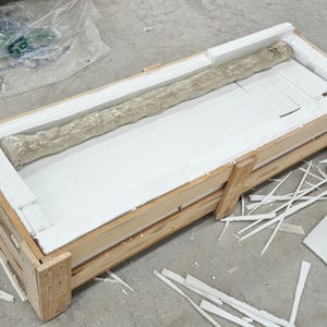 Puede incluir: Un objeto largo y cil&iacute;ndrico envuelto en material protector texturizado de color claro, que descansa dentro de una caja de env&iacute;o rectangular de madera forrada con espuma blanca. La caja est&aacute; construida con listones de madera clara.
