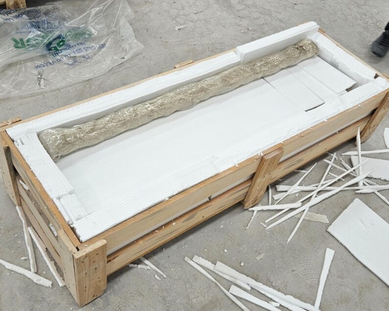 Puede incluir: Un objeto largo y cil&iacute;ndrico envuelto en material protector texturizado de color claro, que descansa dentro de una caja de env&iacute;o rectangular de madera forrada con espuma blanca. La caja est&aacute; construida con listones de madera clara.