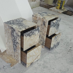 May include: Two marble nightstands with open drawers. The nightstands have a beige and gray veined pattern. The drawers are made of wood. The nightstands are rectangular and have a modern design.