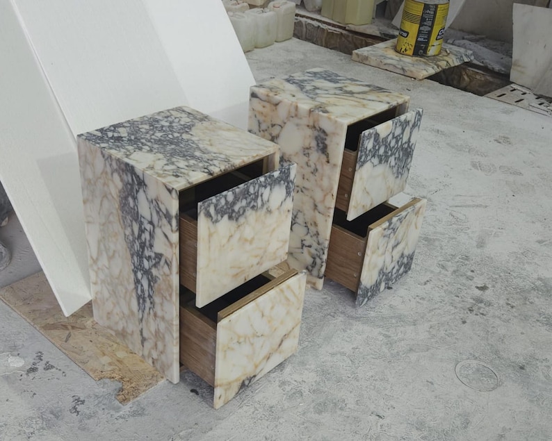May include: Two marble nightstands with open drawers. The nightstands have a beige and gray veined pattern. The drawers are made of wood. The nightstands are rectangular and have a modern design.