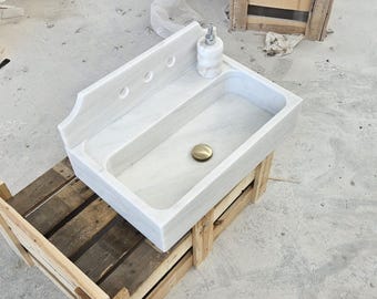 May include: A rectangular white marble sink featuring a high backsplash with three circular faucet holes. The basin includes a centred circular brass drain cover and a matching white marble soap dispenser resting on the top ledge.