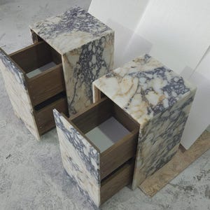 May include: Two marble-patterned nightstands with open drawers. The nightstands have a light beige and gray veined pattern. The drawers are made of wood. The nightstands are cube-shaped and have a modern design.