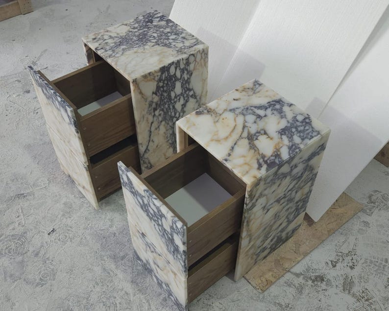May include: Two marble-patterned nightstands with open drawers. The nightstands have a light beige and gray veined pattern. The drawers are made of wood. The nightstands are cube-shaped and have a modern design.