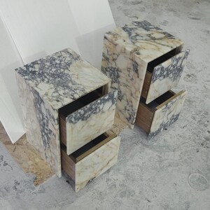 May include: Two marble nightstands with open drawers. The nightstands have a cream and gray veined pattern. The drawers are made of wood and have a dark interior. The nightstands are on a gray concrete floor.