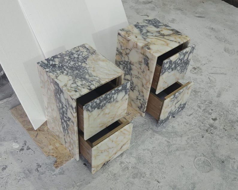 May include: Two marble nightstands with open drawers. The nightstands have a cream and gray veined pattern. The drawers are made of wood and have a dark interior. The nightstands are on a gray concrete floor.