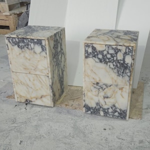 May include: Two marble nightstands with a cream and gray veined pattern. Each nightstand has two drawers and a rectangular shape. The nightstands are made of stone and are placed on a light-colored surface.