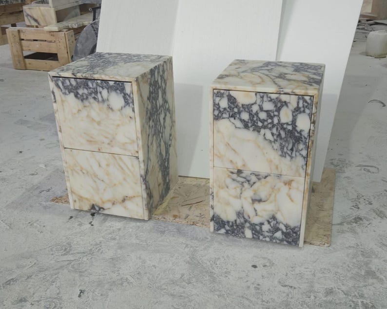 May include: Two marble nightstands with a cream and gray veined pattern. Each nightstand has two drawers and a rectangular shape. The nightstands are made of stone and are placed on a light-colored surface.