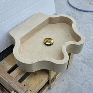 Op de afbeelding: Een beige stenen wastafel met een onregelmatige, organische vorm en een gladde, matte afwerking. De waskom heeft een centraal rond afvoergat voorzien van een gepolijste, reflecterende goudkleurige metalen afvoerplug die contrasteert met de steen.