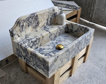 May include: A rectangular marble sink featuring a high back splash and a polished white surface with prominent dark grey and black veining. The basin includes a gold-toned circular drain cover and a white soap dispenser resting on the rear ledge.