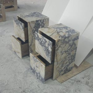 May include: Two marble nightstands with open drawers. The nightstands are cube-shaped with a mix of white, beige, and gray veining. The drawers are open, revealing the wooden interior. The nightstands are on a concrete floor.