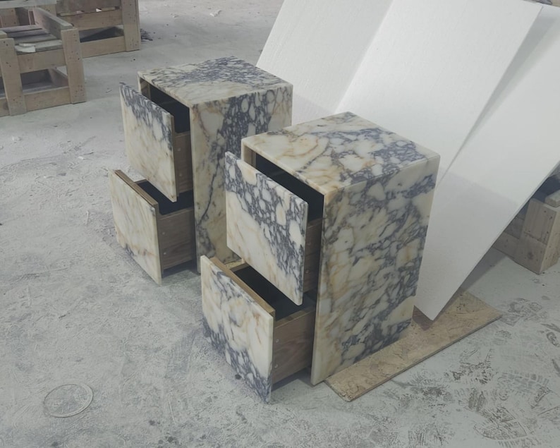 May include: Two marble nightstands with open drawers. The nightstands are cube-shaped with a mix of white, beige, and gray veining. The drawers are open, revealing the wooden interior. The nightstands are on a concrete floor.