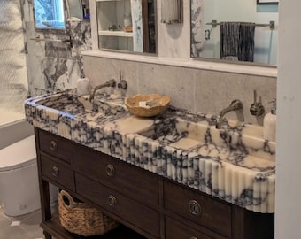 May include: A double vanity featuring a fluted marble countertop with white and dark gray veining, two integrated rectangular sinks, and two silver faucets. The base is a dark brown wooden cabinet with six drawers, round metal pulls, and a woven basket.