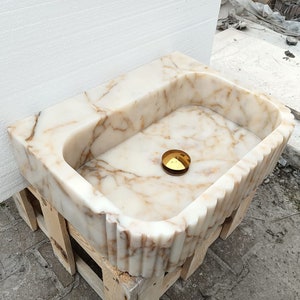 May include: A rectangular marble sink with a scalloped edge and a golden drain cover. The sink has a cream and brown veined pattern. The sink is sitting on a wooden pallet.