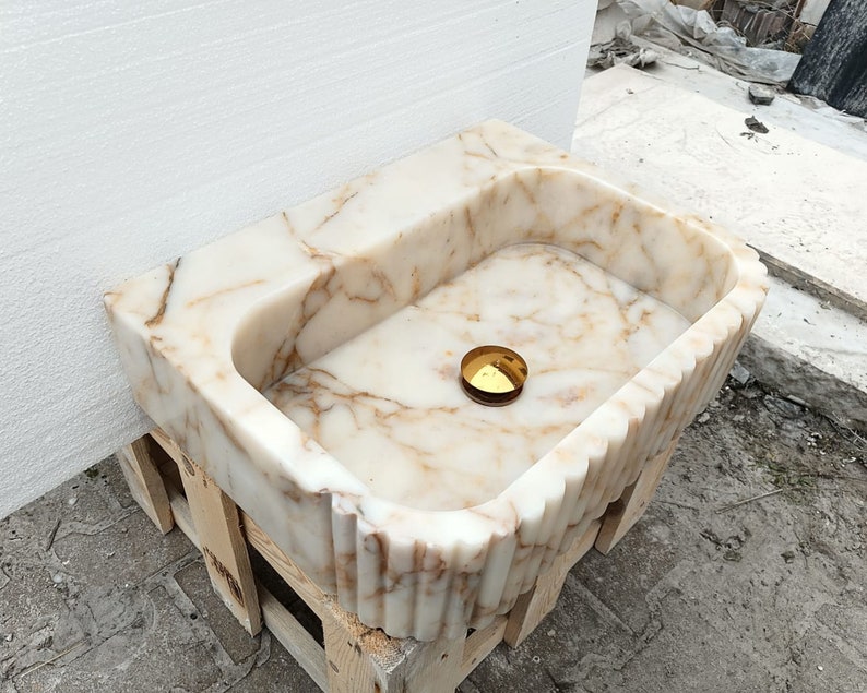 May include: A rectangular marble sink with a scalloped edge and a golden drain cover. The sink has a cream and brown veined pattern. The sink is sitting on a wooden pallet.