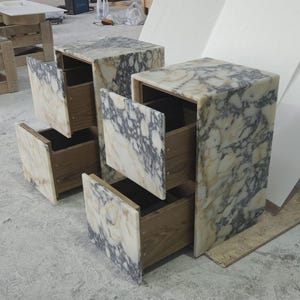 May include: Two marble-topped nightstands with open drawers. The marble has a cream and gray veined pattern. The drawers are made of wood. The nightstands are rectangular and have a modern design.