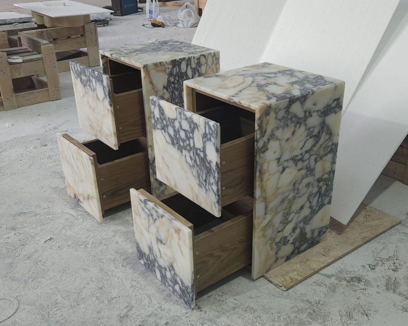 May include: Two marble-topped nightstands with open drawers. The marble has a cream and gray veined pattern. The drawers are made of wood. The nightstands are rectangular and have a modern design.
