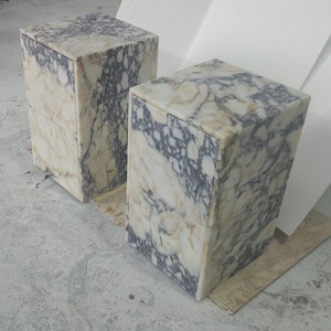 May include: Two marble cube-shaped pedestals with a cream, gray, and black veined pattern. The pedestals are side by side, showcasing the natural stone's unique veining. The cubes are on a light gray surface.