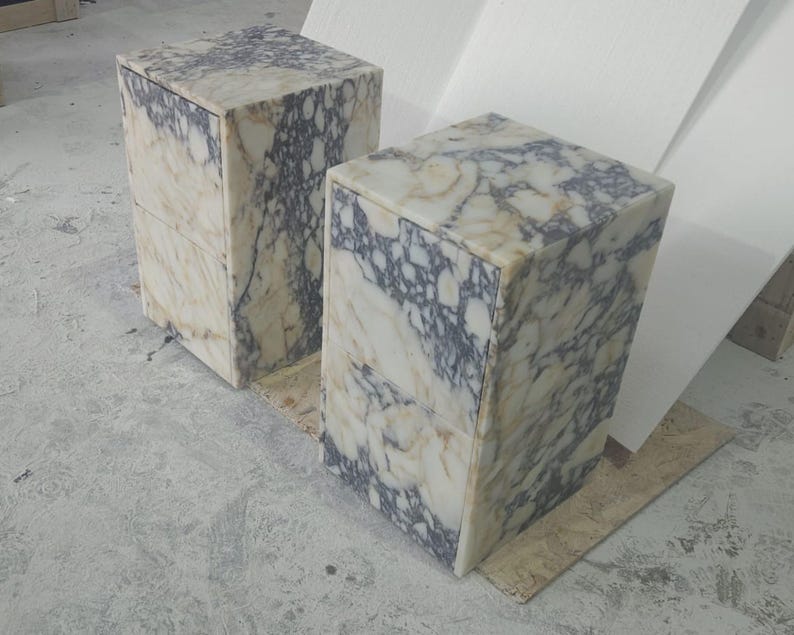 May include: Two marble cube-shaped pedestals with a cream, gray, and black veined pattern. The pedestals are side by side, showcasing the natural stone's unique veining. The cubes are on a light gray surface.