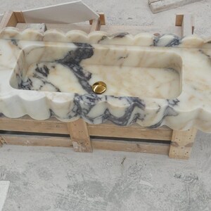 Puede incluir: Un lavabo rectangular de m&aacute;rmol con borde festoneado y vetas en crema, blanco y gris oscuro. La cubeta incluye un tap&oacute;n de desag&uuml;e dorado y un dispensador de jab&oacute;n blanco se encuentra en el lado derecho de la superficie de la encimera.