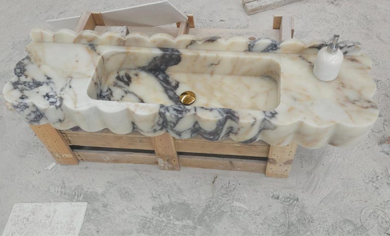 Puede incluir: Un lavabo rectangular de m&aacute;rmol con borde festoneado y vetas en crema, blanco y gris oscuro. La cubeta incluye un tap&oacute;n de desag&uuml;e dorado y un dispensador de jab&oacute;n blanco se encuentra en el lado derecho de la superficie de la encimera.