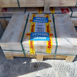 May include: A wooden pallet wrapped in clear plastic, secured with green straps. Yellow and blue DHL tape with the text "Mikro İhracat" is affixed to the package. The pallet is stacked with other similar packages.