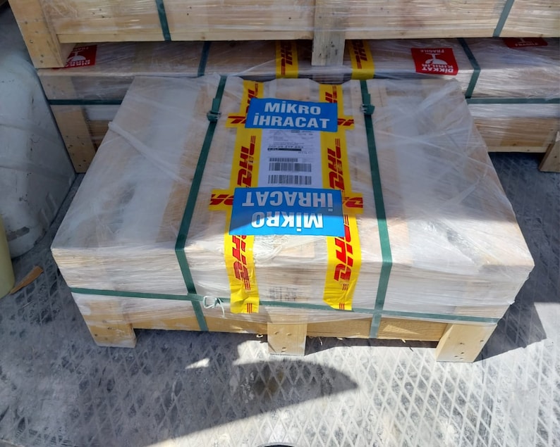 May include: A wooden pallet wrapped in clear plastic, secured with green straps. Yellow and blue DHL tape with the text "Mikro İhracat" is affixed to the package. The pallet is stacked with other similar packages.