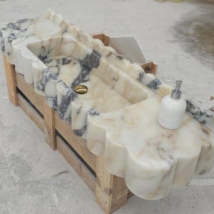 Puede incluir: Un lavabo rectangular de m&aacute;rmol con borde festoneado y vetas blancas y grises. La cubeta incluye un desag&uuml;e de tono dorado y un dispensador de jab&oacute;n blanco se encuentra sobre la superficie plana de la estructura de piedra del mostrador.
