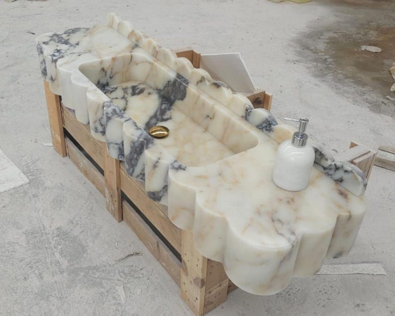 Puede incluir: Un lavabo rectangular de m&aacute;rmol con borde festoneado y vetas blancas y grises. La cubeta incluye un desag&uuml;e de tono dorado y un dispensador de jab&oacute;n blanco se encuentra sobre la superficie plana de la estructura de piedra del mostrador.