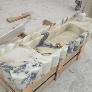 Puede incluir: Un lavabo rectangular de m&aacute;rmol con borde festoneado y vetas blancas y grises. La cubeta incluye una tapa de desag&uuml;e dorada pulida y un dispensador de jab&oacute;n blanco se asienta en la esquina de la superficie de piedra, mostrando patrones naturales.