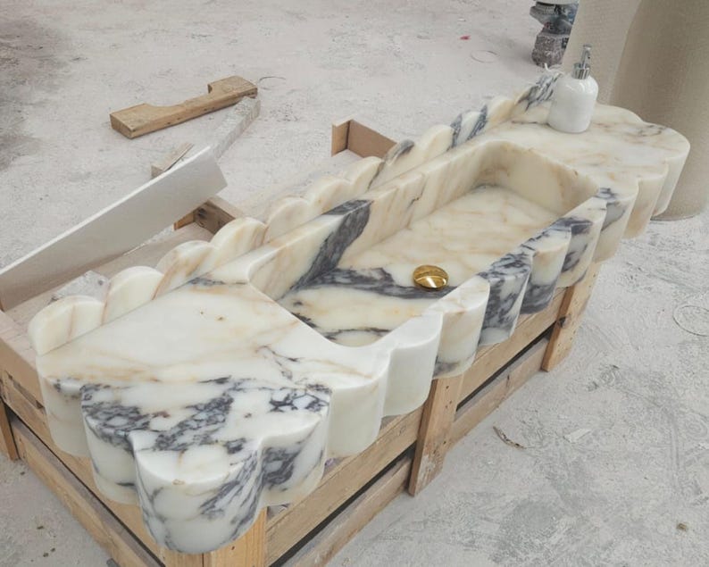 Puede incluir: Un lavabo rectangular de m&aacute;rmol con borde festoneado y vetas blancas y grises. La cubeta incluye una tapa de desag&uuml;e dorada pulida y un dispensador de jab&oacute;n blanco se asienta en la esquina de la superficie de piedra, mostrando patrones naturales.