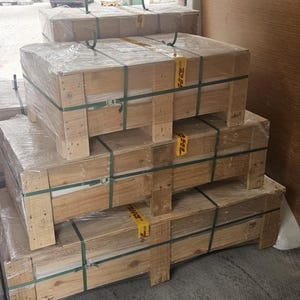 May include: Stacked wooden crates wrapped in clear plastic and secured with green straps. The crates are rectangular and appear to be ready for transport. Yellow DHL labels are visible on some of the crates.
