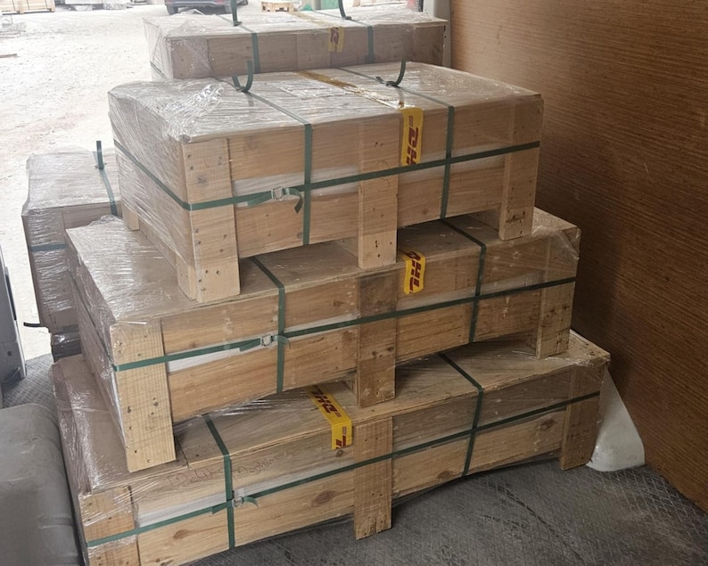 May include: Stacked wooden crates wrapped in clear plastic and secured with green straps. The crates are rectangular and appear to be ready for transport. Yellow DHL labels are visible on some of the crates.