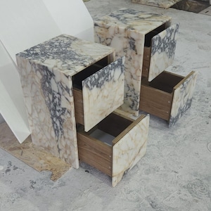 May include: Two marble-patterned nightstands with open drawers. The nightstands have a cream and gray color scheme, with wooden drawer interiors. The drawers are partially open, revealing the interior space. The nightstands are cube-shaped.