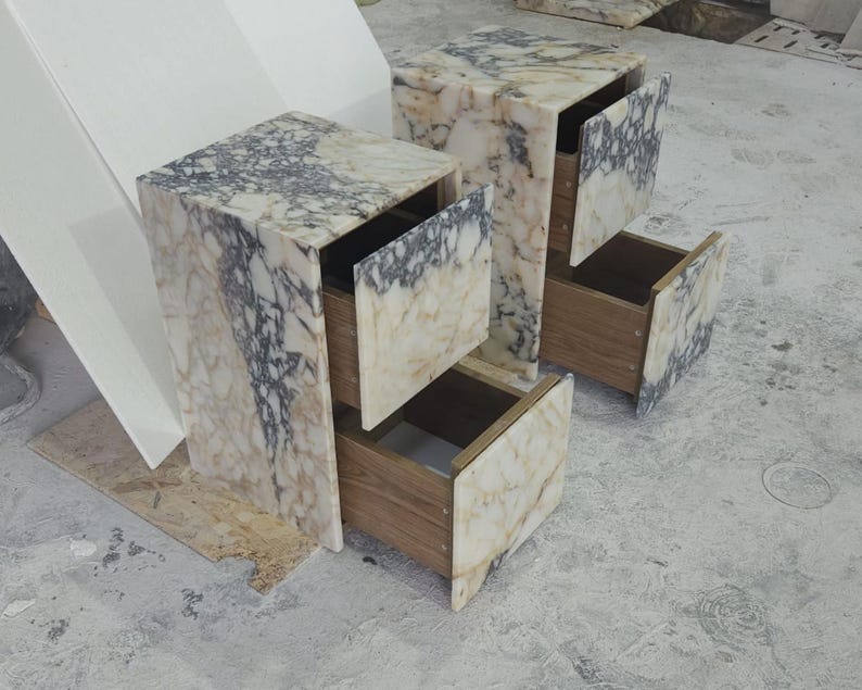 May include: Two marble-patterned nightstands with open drawers. The nightstands have a cream and gray color scheme, with wooden drawer interiors. The drawers are partially open, revealing the interior space. The nightstands are cube-shaped.