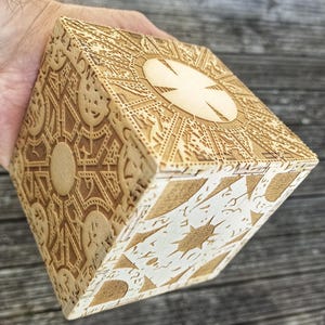 May include: A wooden puzzle box with intricate carvings and a circular design on the top. The box is made of light brown wood and has a natural finish.
