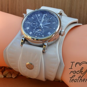 May include: A silver and white wristwatch with a blue face and multiple complications, including a moon phase indicator, day of the week, and date. The watch is on a white leather strap with a buckle fastening.