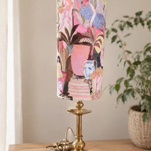 May include: A table lamp with a cylindrical fabric shade featuring a colorful, painterly illustration of figures, plants, and vases in shades of pink, blue, and brown. The lamp has a polished brass base with a slender stem and a circular foot.