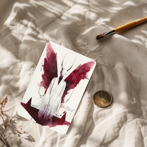 Könnte beinhalten: Eine Tarotkarte mit einer Aquarellillustration einer Figur mit roten Flügeln und dem Text "The Devil". Die Karte liegt auf einer weißen Oberfläche, mit einer Goldmünze und einem Pinsel in der Nähe.